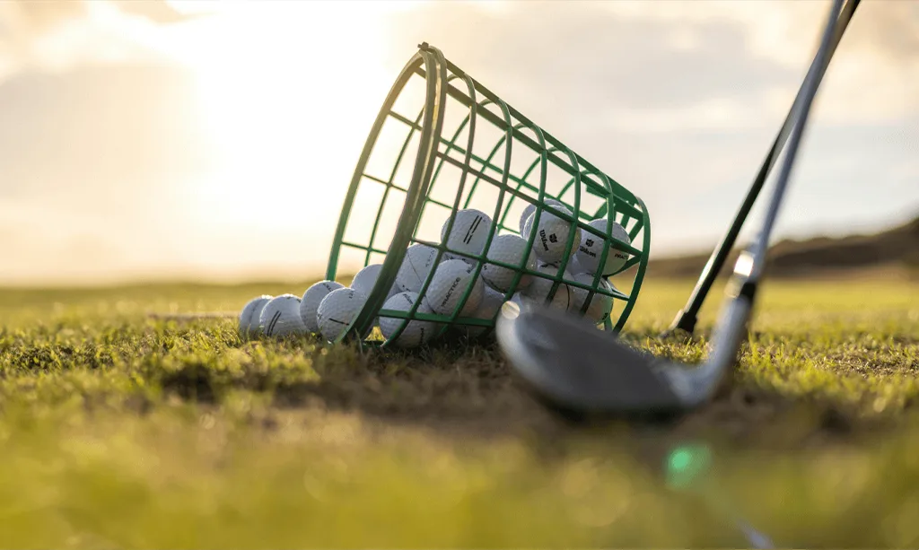 Un panier de balles renversé sur l’herbe d’un champ de pratique, au soleil couchant.